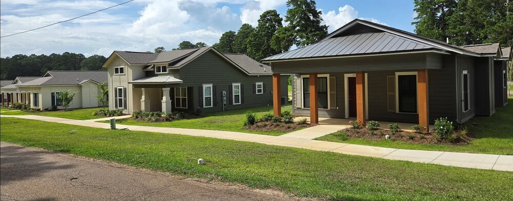 A dark grey house with cedar pillars, next to a dark green house with white pillars with a sidewalk in front of them.